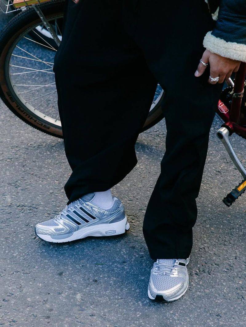 Close-up of silver Adistar Control 5 sneakers with black trousers, next to a bicycle.